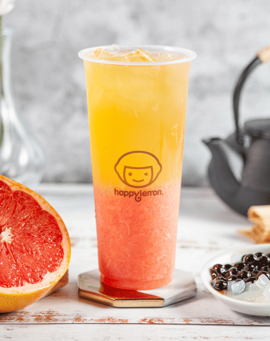 A tall plastic cup of layered yellow and pink iced drink sits on a coaster, with grapefruit, a bowl of black tapioca pearls, and a teapot in the background. The cup has a "happy lemon" logo on it.