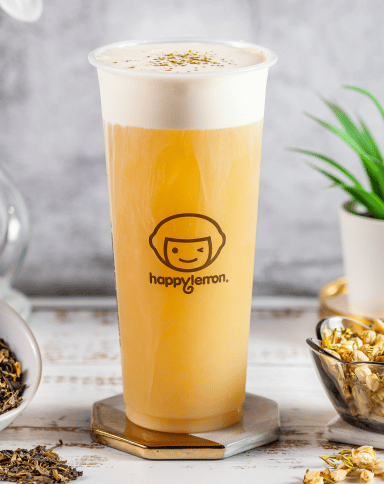 A tall plastic cup of tea with a frothy top sits on a coaster. The cup has the "happy lemon" logo on it. The background includes a small plant, bowls of tea leaves, and a light gray textured backdrop.