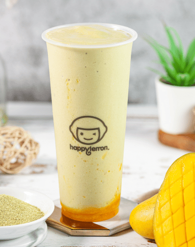 A tall plastic cup with a light yellow smoothie, featuring a layer of orange syrup at the bottom. The cup has a “happy lemon” logo. A sliced mango, a bowl of powder, and a small potted plant are nearby.