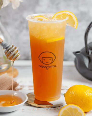 A tall plastic cup of iced tea with lemon slices, garnished with a lemon wedge. A honey dipper drips honey into a small dish beside a whole lemon, and a teapot is partially visible in the background.
