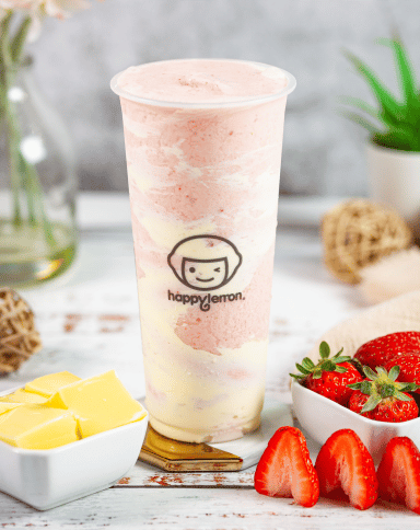 A tall cup of swirled pink and yellow blended drink, labeled “happy lemon,” is placed on a white surface with cubes of yellow fruit, whole and sliced strawberries, and decorative items in the background.