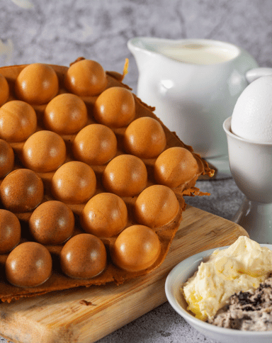 A bubble waffle sits on a wooden board next to a bowl of cream and cookies, a whole egg in an egg cup, and a white pitcher of milk, against a gray textured background.