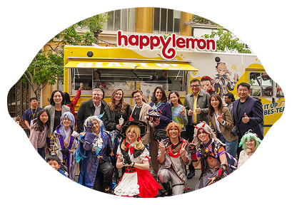 A group of people, including several in colorful cosplay costumes, pose in front of a yellow Happy Lemon food truck. Some people are standing while others crouch in front, all smiling for the photo.