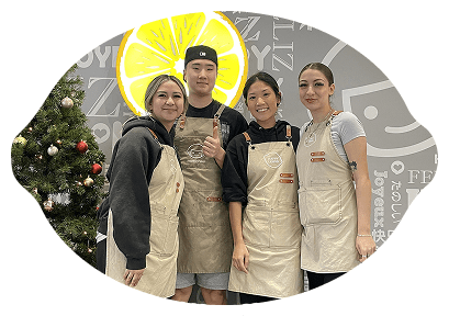 Four people wearing beige aprons stand together and smile in front of a decorated Christmas tree and a wall with text designs. A large yellow lemon graphic is on the wall behind them.
