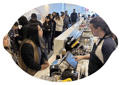 A group of people stands in line at a fast-casual restaurant counter, where an employee in an apron assists a customer using a touchscreen register. Toppings and ingredients are visible on the counter.