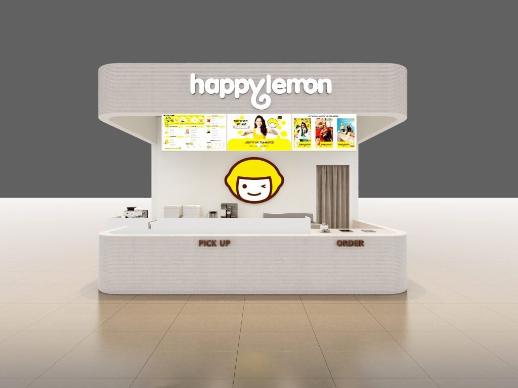 A white kiosk with the sign "happylemon" at the top, menu boards above the counter, and a smiling cartoon face logo in the center. The counter has labeled "PICK UP" and "ORDER" sections.