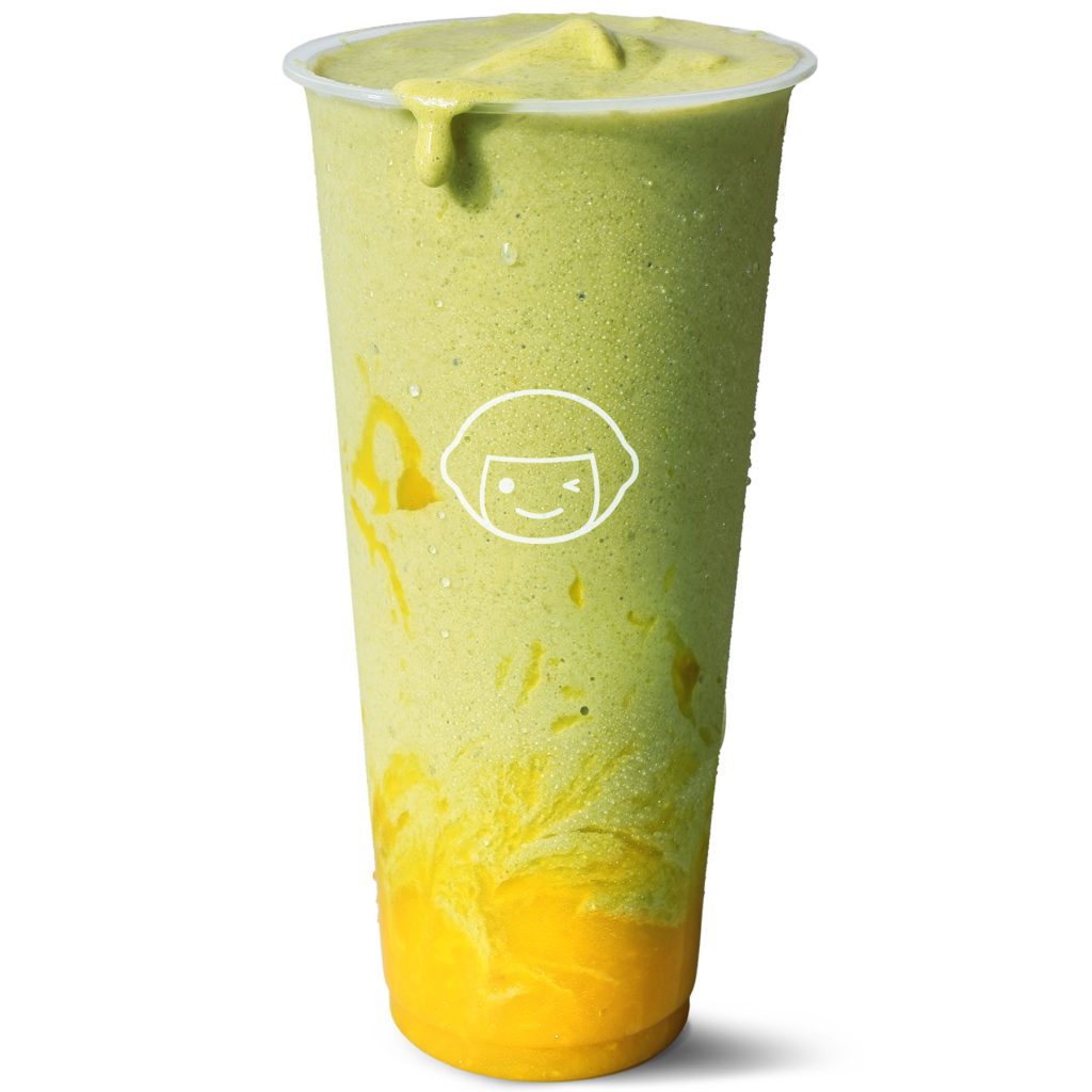 A tall clear cup filled with a green smoothie and bright yellow puree swirled at the bottom, with a small smiley face logo on the front of the cup and some smoothie dripping over the rim.