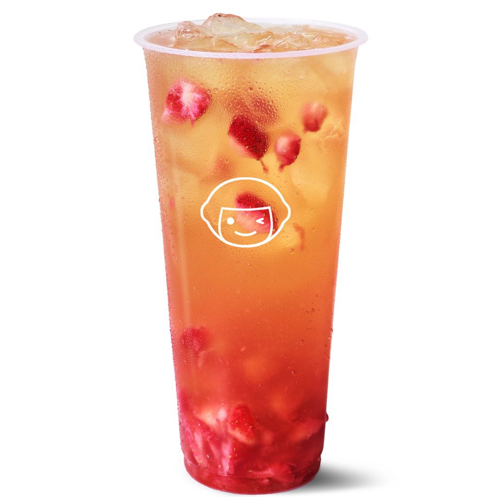 A clear plastic cup filled with an iced, orange-yellow drink containing pieces of red fruit, likely strawberries. The cup features a white cartoon face logo on the front.