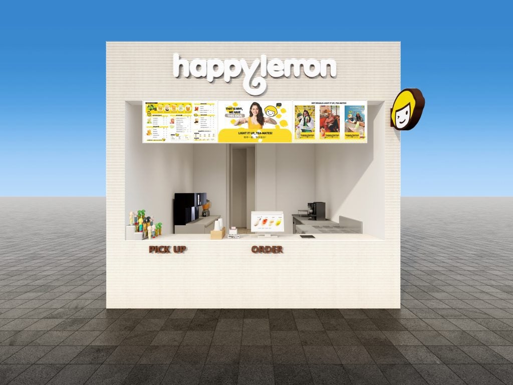A front view of a Happy Lemon kiosk with a white exterior, menu boards above the counter, and labeled "Pick Up" and "Order" sections. The logo and a cartoon lemon face sign are visible. The background is clear and blue.