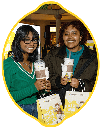 Two people smile at the camera while holding drinks topped with whipped cream and straws. Both are holding shopping bags that read "Blast of Flavor" in a decorated indoor setting.