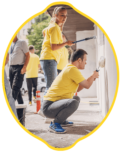 Two people dressed in yellow shirts are painting an outdoor wall white. One person is standing and the other is crouched down, both using paint rollers. Other individuals are visible in the background.