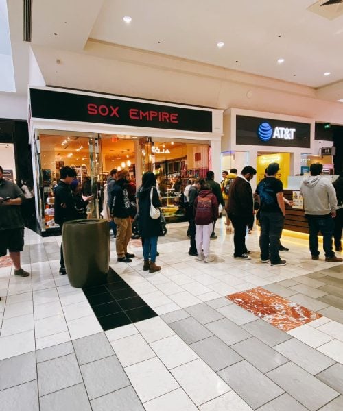 People stand in two lines inside a shopping mall. The lines form in front of different stores and kiosks, including one labeled "SOX EMPIRE." The floor is tiled, and the area is brightly lit.