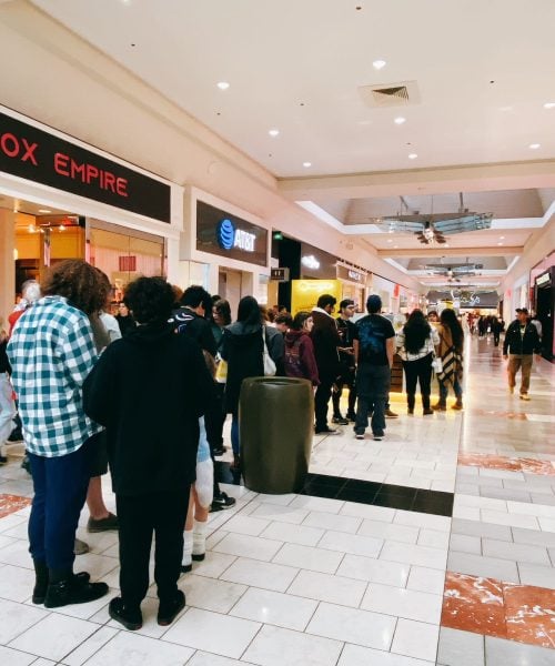 A group of people stand in a line inside a shopping mall, near various store fronts including Box Empire, AT&T, and others. Shoppers walk along the mall’s tiled floor under bright ceiling lights.