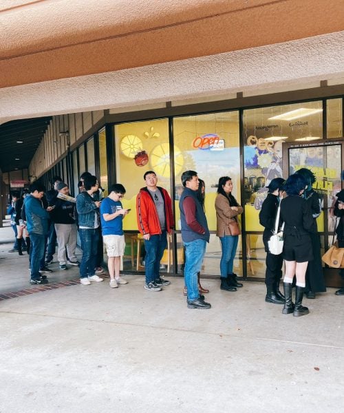 A long line of people, some in casual clothing and others in cosplay, waits outside a storefront with large windows and an "open" sign during the day. The building has a covered walkway and a potted plant nearby.