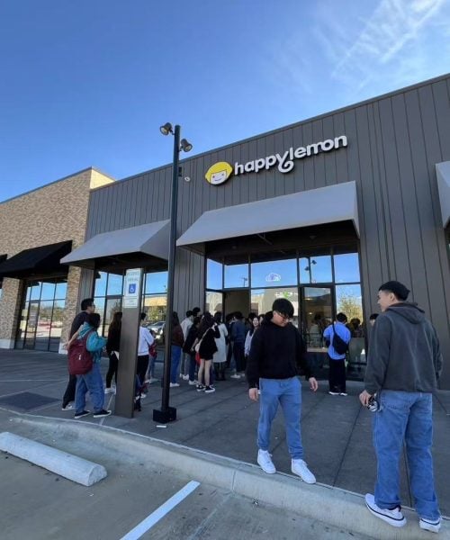 A group of people stand in line outside a Happy Lemon store in a shopping plaza on a sunny day. Some are gathered near the entrance while others stand nearby. The neighboring store, Chinchikurin, is also visible.