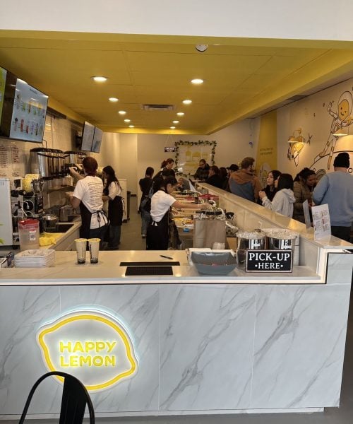 People stand in line at a brightly lit bubble tea shop with a yellow ceiling. Staff prepare drinks behind a counter with a neon "Happy Lemon" sign and a "Pick-Up Here" area. Menu screens are visible on the left wall.