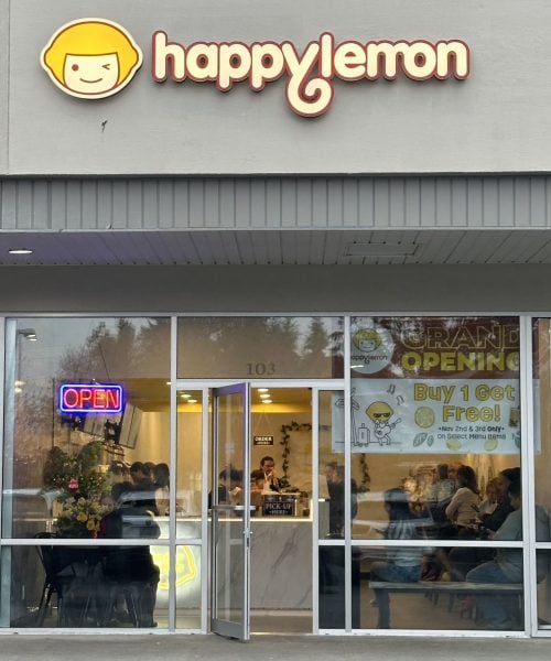 A Happy Lemon storefront with a yellow logo, a large window, and an "OPEN" sign. People are seated inside. A banner advertises a grand opening and a "Buy 1 Get 1 Free" promotion.