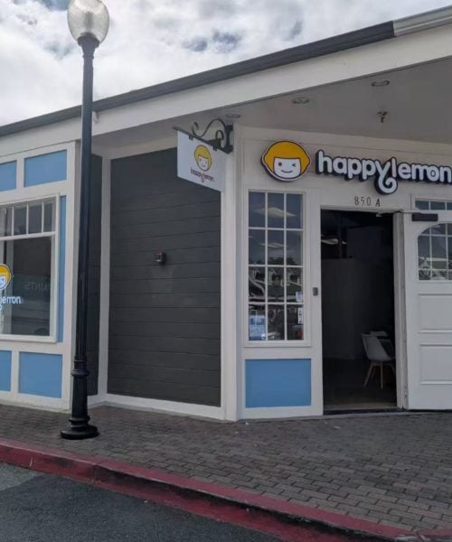 A storefront with a sign reading "happy lemon" and a logo featuring a smiling face with yellow hair. The building has blue and white trim, large windows, and an open door. A sidewalk and lamp post are in front.