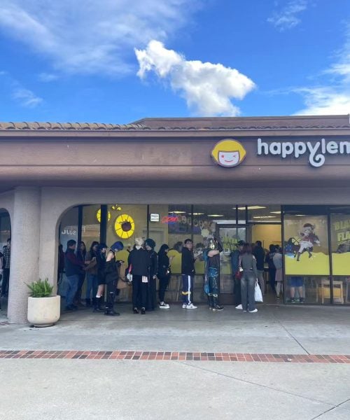 People stand in a long line outside a Happy Lemon shop in a shopping plaza on a sunny day. The store has large windows with bright yellow signs and a logo above the entrance.