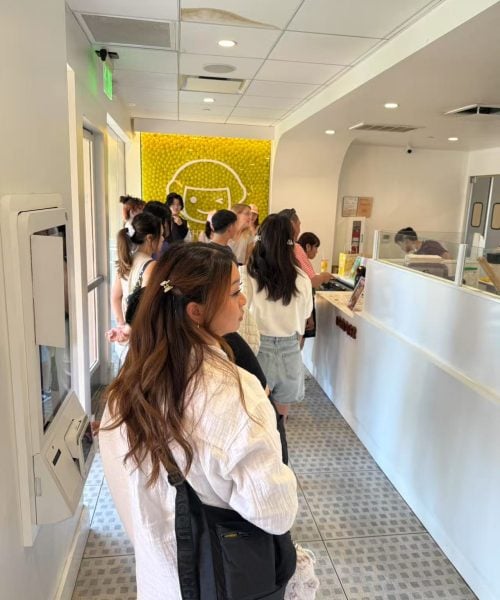 A line of people waits inside a brightly lit dessert shop with white walls and a tiled floor. Some customers are looking at the counter staff. A wall decorated with yellow accents and a cartoon face is in the background.