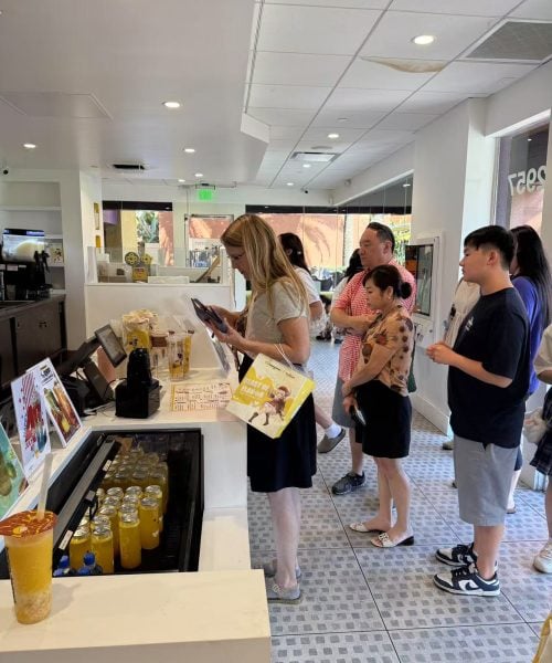 A group of people stands in line inside a bright, modern cafe. The woman at the counter is ordering while others wait behind her. Bottled drinks and menu displays are visible on the counter.