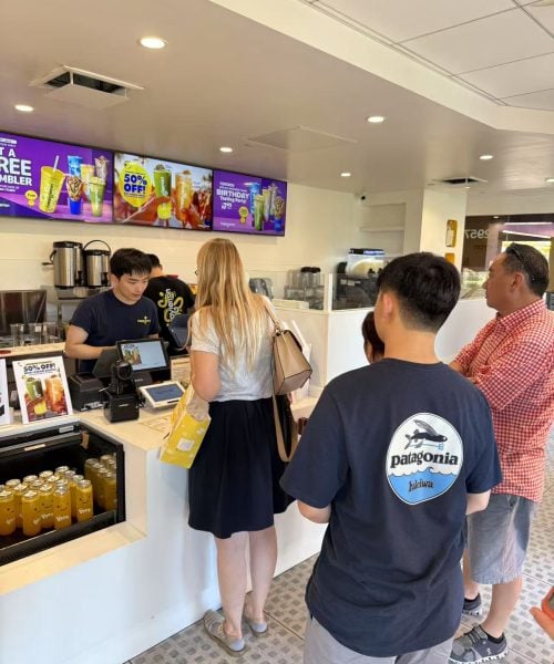 Customers stand in line at a counter inside a brightly lit bubble tea shop. A staff member takes an order, and several promotional drinks are displayed on screens and the counter.