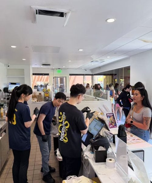 Employees behind a counter take orders and prepare drinks for customers at a busy café. Customers wait in line inside the bright shop, with various drink supplies and equipment visible on the counter.