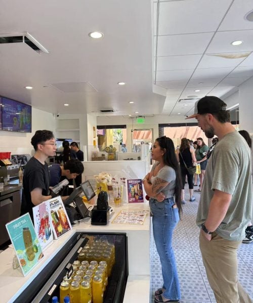 Two customers stand at a counter speaking with an employee in a bright, modern cafe. Bottled drinks and fruit tea advertisements are visible on the counter, and other people are in the background.
