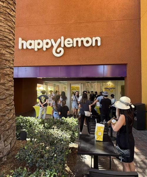 People stand in line outside a Happy Lemon store with a purple awning. Some customers wait at tables in front of the shop, and others gather near the entrance on a sunny day. A palm tree is visible on the left.