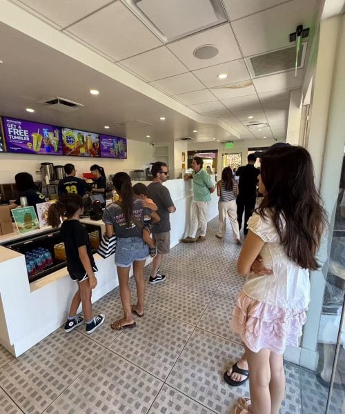 People stand in line inside a brightly lit shop, waiting to order at the counter where employees are serving customers. Digital menu boards and promotional signs are visible above the counter.