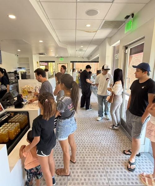 A group of people stand in line inside a bright, modern cafe. Some are ordering at the counter while others wait. Several drinks are visible on the counter, and sunlight comes in through the glass door.