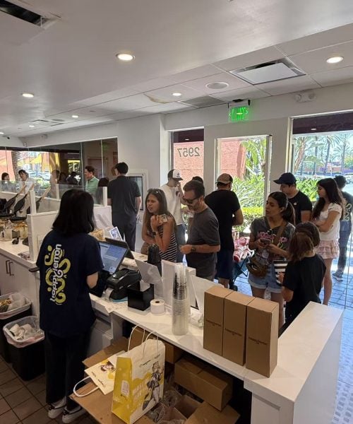 Customers stand in line at a counter inside a casual restaurant or café. A staff member is taking an order at the register, and various items and packaging are visible on the counter. The atmosphere is busy.