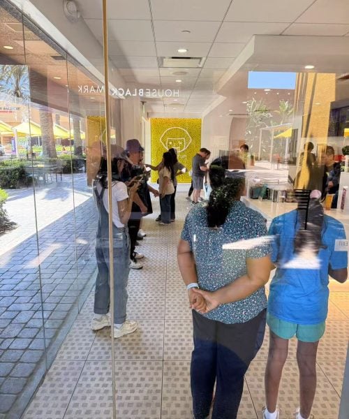 People stand in line inside a store with yellow decor, seen through a glass window. Some people are outside, visible through reflections. The scene is bright with sunlight and outdoor seating is visible.