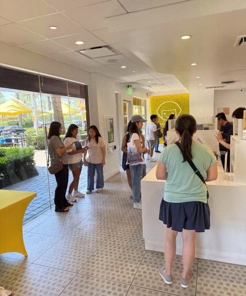 Several people stand in line or wait inside a bright, modern shop with white walls and yellow accents. Large windows reveal outdoor seating with yellow umbrellas. Some people appear to be reading menus.