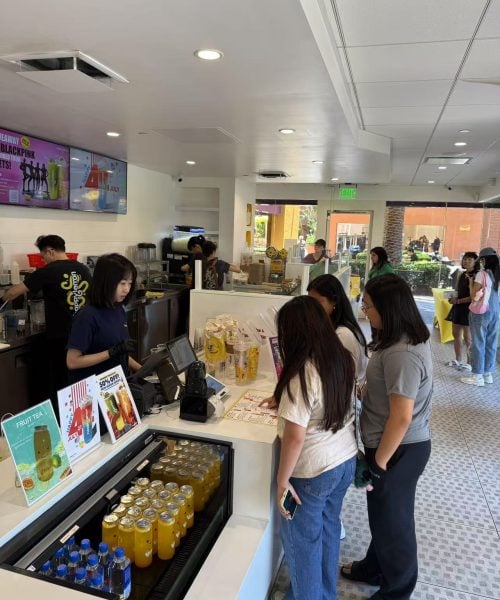 Several people stand in line at a counter in a modern cafe or drink shop. A worker takes orders at the register. Bottled drinks and promotional displays are on the counter. Other customers are visible in the background.