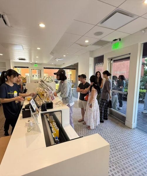 A group of customers waits in line inside a modern, brightly lit cafe. A staff member stands behind the counter, ready to take orders. Additional customers stand outside, visible through the glass door.