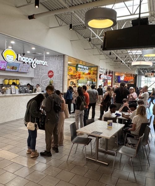 A food court with people standing in line at a counter labeled "happy lemon," others walking or sitting at tables, and various restaurant signs visible in the background. The space has high ceilings and natural light.