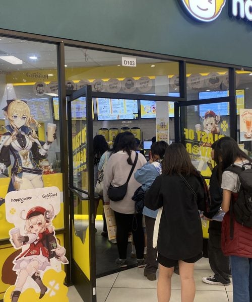 A group of people wait in line inside a brightly lit Happy Lemon store. Cardboard cutouts of anime characters are displayed at the entrance, and digital menus are visible above the counter.