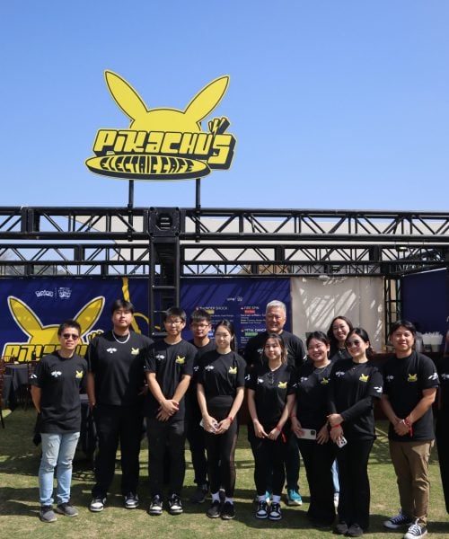 A group of people wearing matching black Pikachu-themed shirts stand in front of a booth with a large yellow Pikachu sign on top, under a clear blue sky.