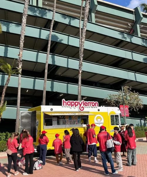 A group of people, most wearing red shirts, stand in line at a yellow Happy Lemon food truck parked outside a multi-level parking structure with several tall palm trees nearby.