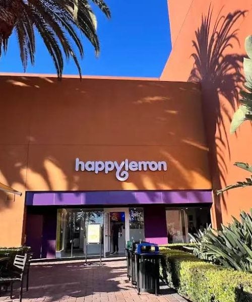 The front entrance of a Happy Lemon store with orange walls, purple accents, and a large palm tree casting a shadow on the building. There are outdoor tables and chairs and some greenery in the foreground.